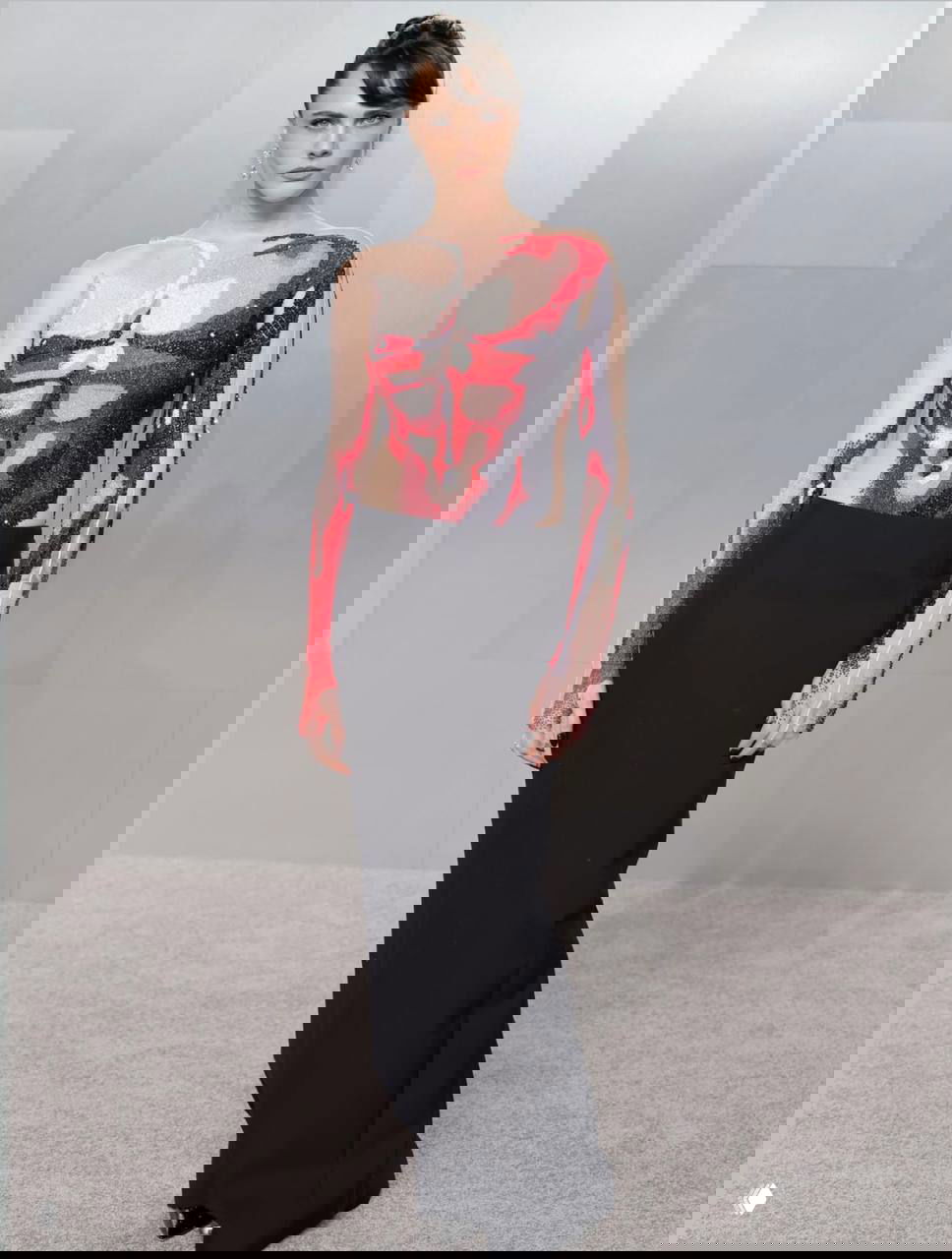 Женщина в чёрной длинной юбке и прозрачном топе с анатомическим принтом, собранные волосы и акцент на украшениях на фоне серого ковра.