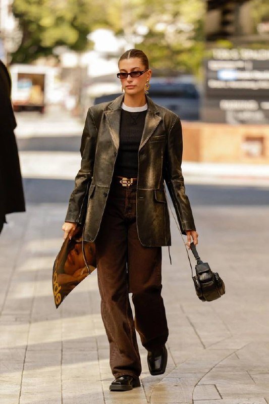 Шоколадный деним в монохромном образе: девушка в тёмно‑коричневых джинсах и кожаном пиджаке идёт по тротуару с маленькой сумкой.