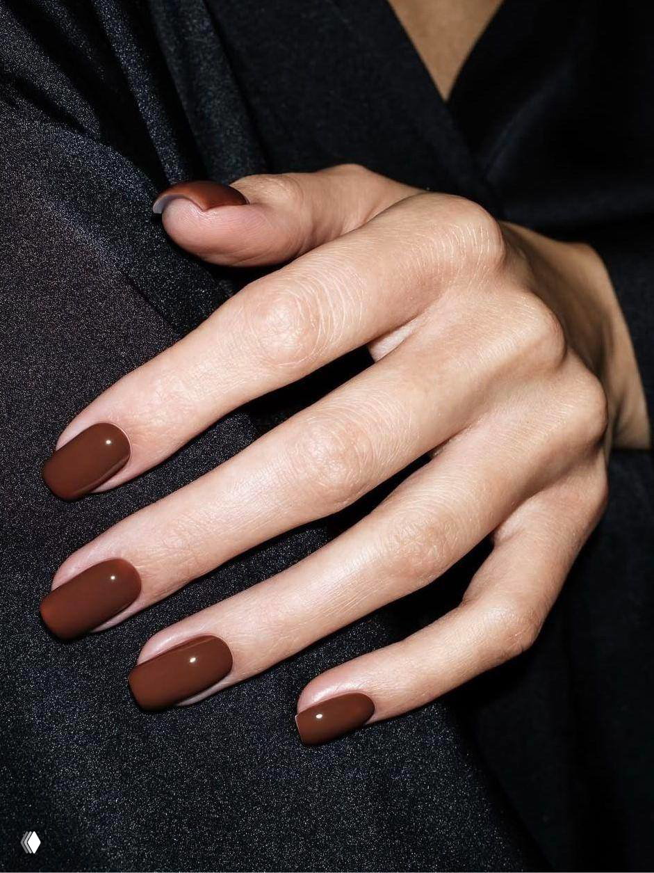 Рука с глянцевым тёмно-коричневым маникюром: ровный овальный силуэт ногтей, видна текстура кожи и минимализм образа.