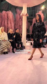 Модель в черном платье идет мимо первого ряда на показе Chanel на фоне больших грибов и розовых театральных декораций, первые зрители в кадре