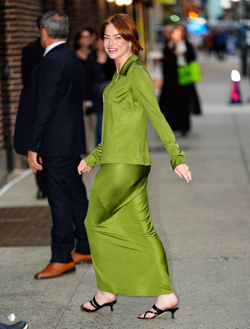 Актриса в зелёном облегающем комплекте шагает по улице, взгляд в сторону, на заднем плане прохожие и городской пейзаж.