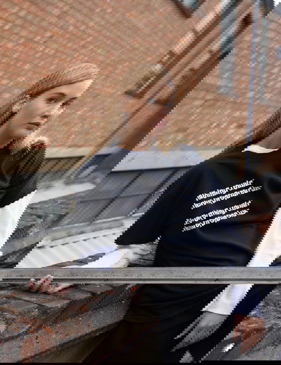 Вертикальный снимок на улице: женщина в свободной футболке и многослойном образе на фоне кирпичной стены и урбанистического пейзажа.