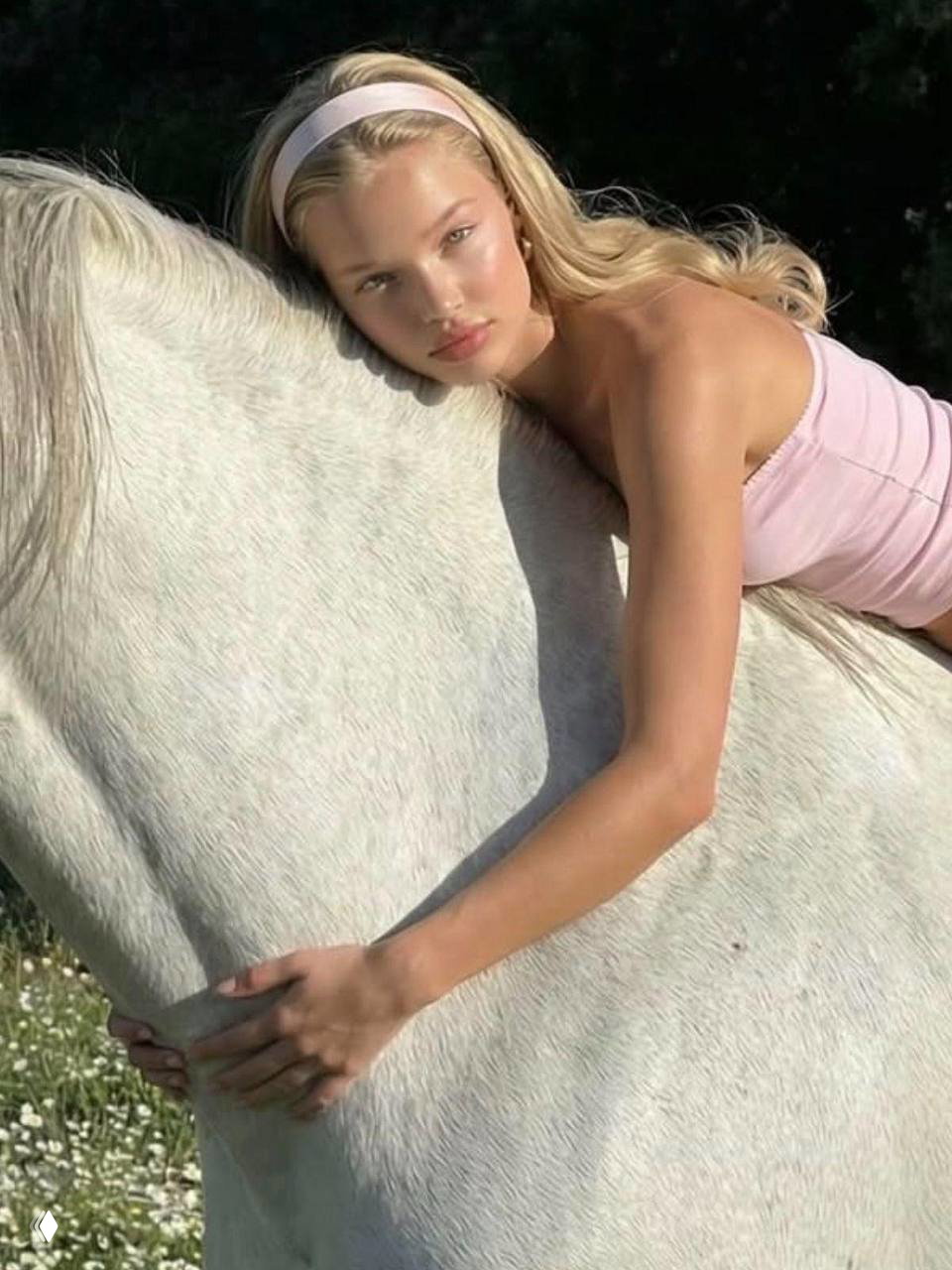 Модель в светлом топе лежит на белой лошади в поле; кадр подчёркивает загар, естественное сияние кожи и мягкую летнюю атмосферу на природе.