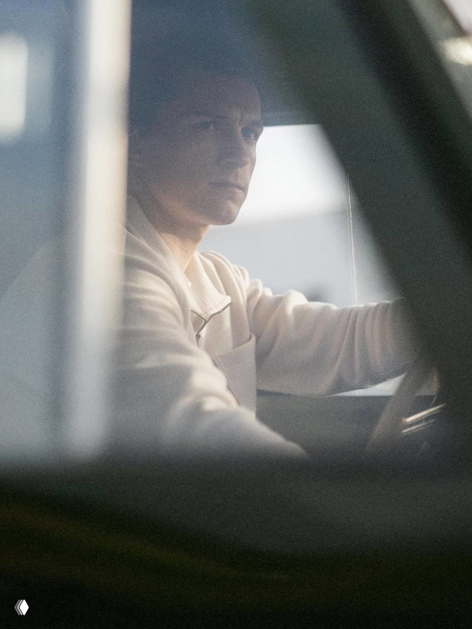 Кадр крупным планом: мужчина за рулём автомобиля в лёгкой спортивной куртке, интерьер машины частично заслоняет композицию кадра, подчёркнуто кинематично.