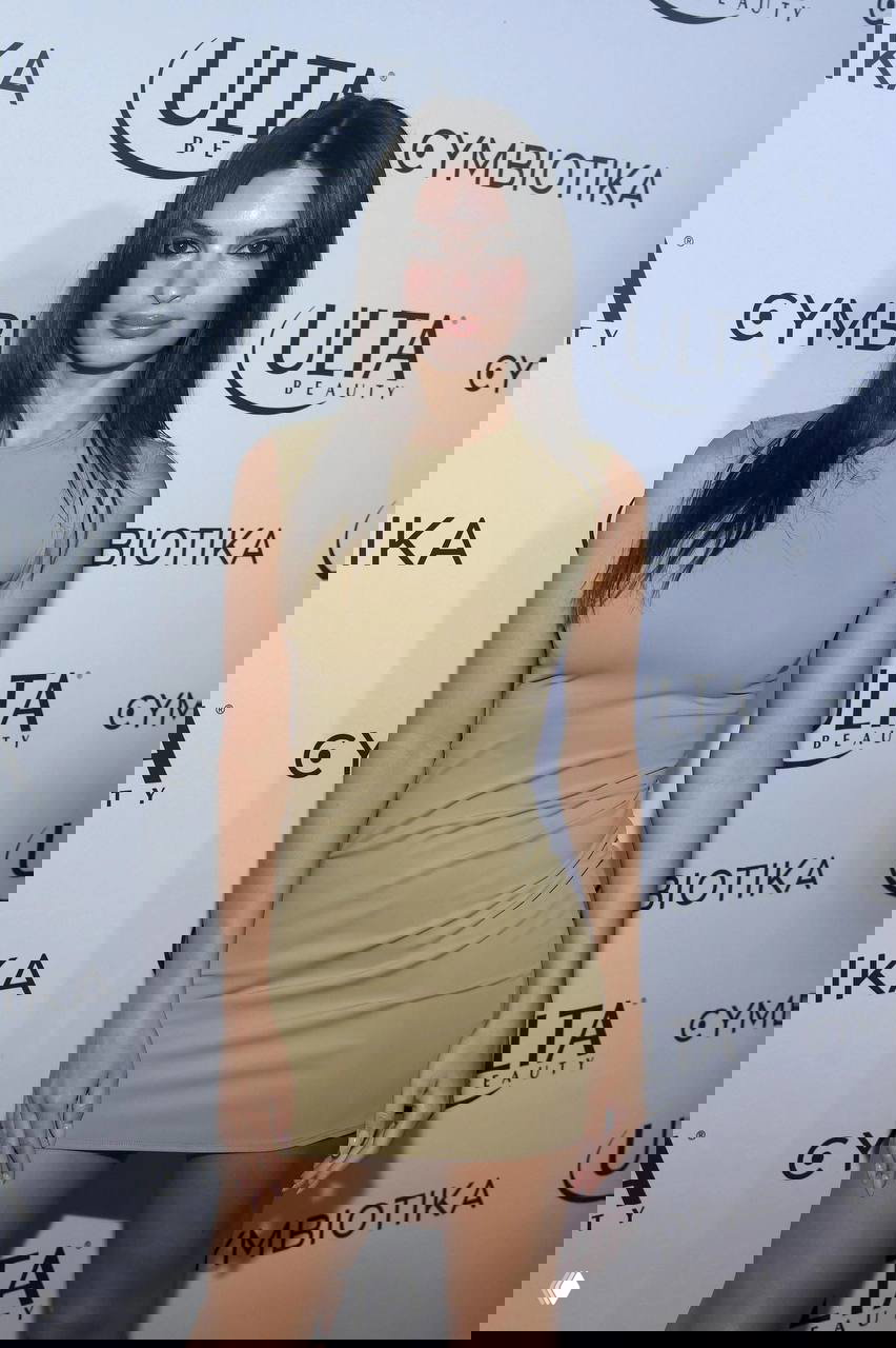 Образ на красной дорожке в облегающем бежевом мини‑платье на фоне брендированных стендов, вертикальный кадр, акцент на посадке платья