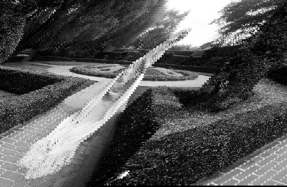 Черно-белая широкоугольная фотография: женщина в струящемся вечернем платье идёт по вымощенной дорожке в ухоженном саду для глянца.
