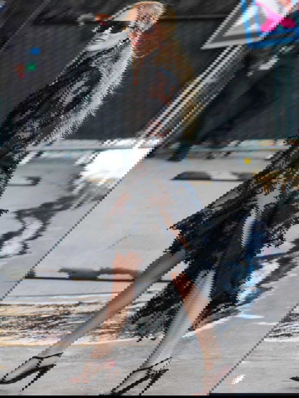 Марго Робби в полупрозрачном кружевном чёрном платье идёт по улице в солнечный день, на ней солнцезащитные очки и открытые босоножки.