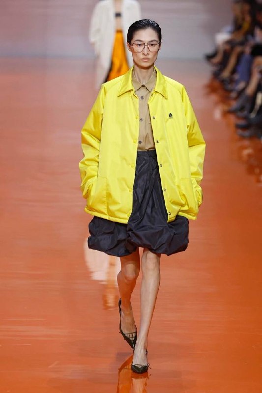 Подиумная модель в ярко-жёлтой ветровке, с коричневой рубашкой и объёмной юбкой — показ, где спортивная куртка выступает главным акцентом.