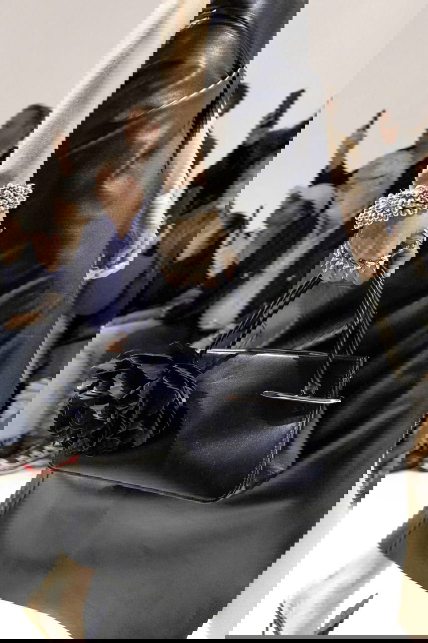 Кадр с подиума: модель в кожаном платье держит сумку, на запястье заметны крупные браслеты-каффы и массивные кольца — динамика показа.