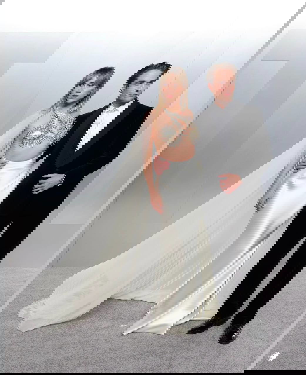 Дуэт: женщина в белом атласном платье с длинной драпированной накидкой и мужчина в чёрном смокинге позируют на фоне логотипа Vanity Fair.