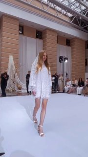 Модель в коротком белом воздушном свадебном платье на подиуме показа EDEM; интерьер с колоннами и зрителями на заднем плане, показ коллекции Demi-Couture.