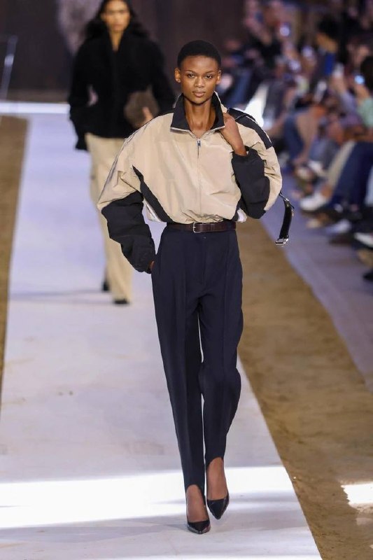 Модель на подиуме в бежево-чёрной ветровке с высокой талией и строгими брюками, элегантные туфли и минималистичный силуэт — подиумный вариант ветровки.