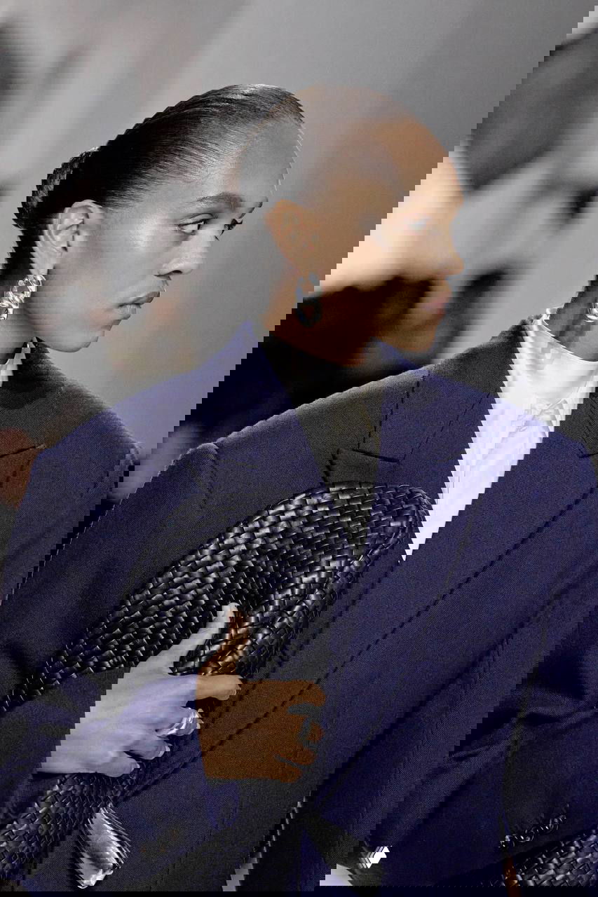 Профиль модели со сверкающими серебристыми серьгами и плетёным клатчем; строгий образ дополнен упрощённым костюмом и гладкой причёской.