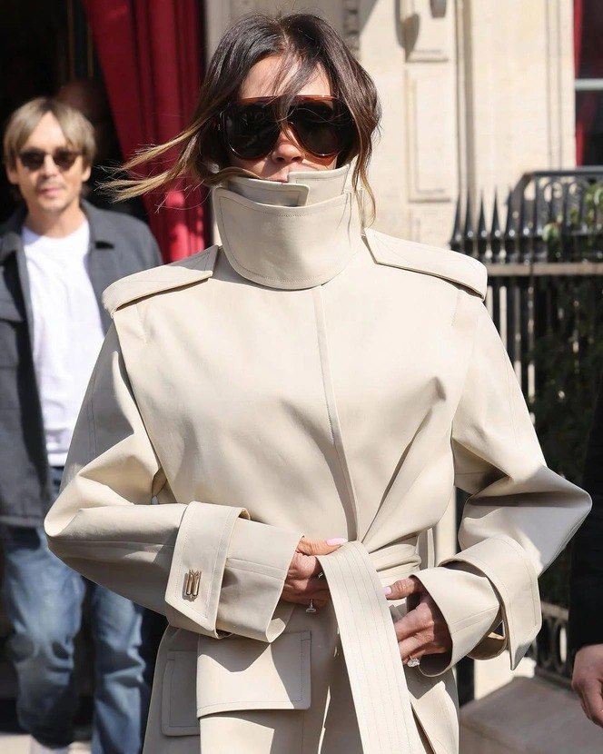 Женщина в светлом тренче с высоким воротником, волосы частично убраны внутрь воротника, крупные солнцезащитные очки и аксессуары