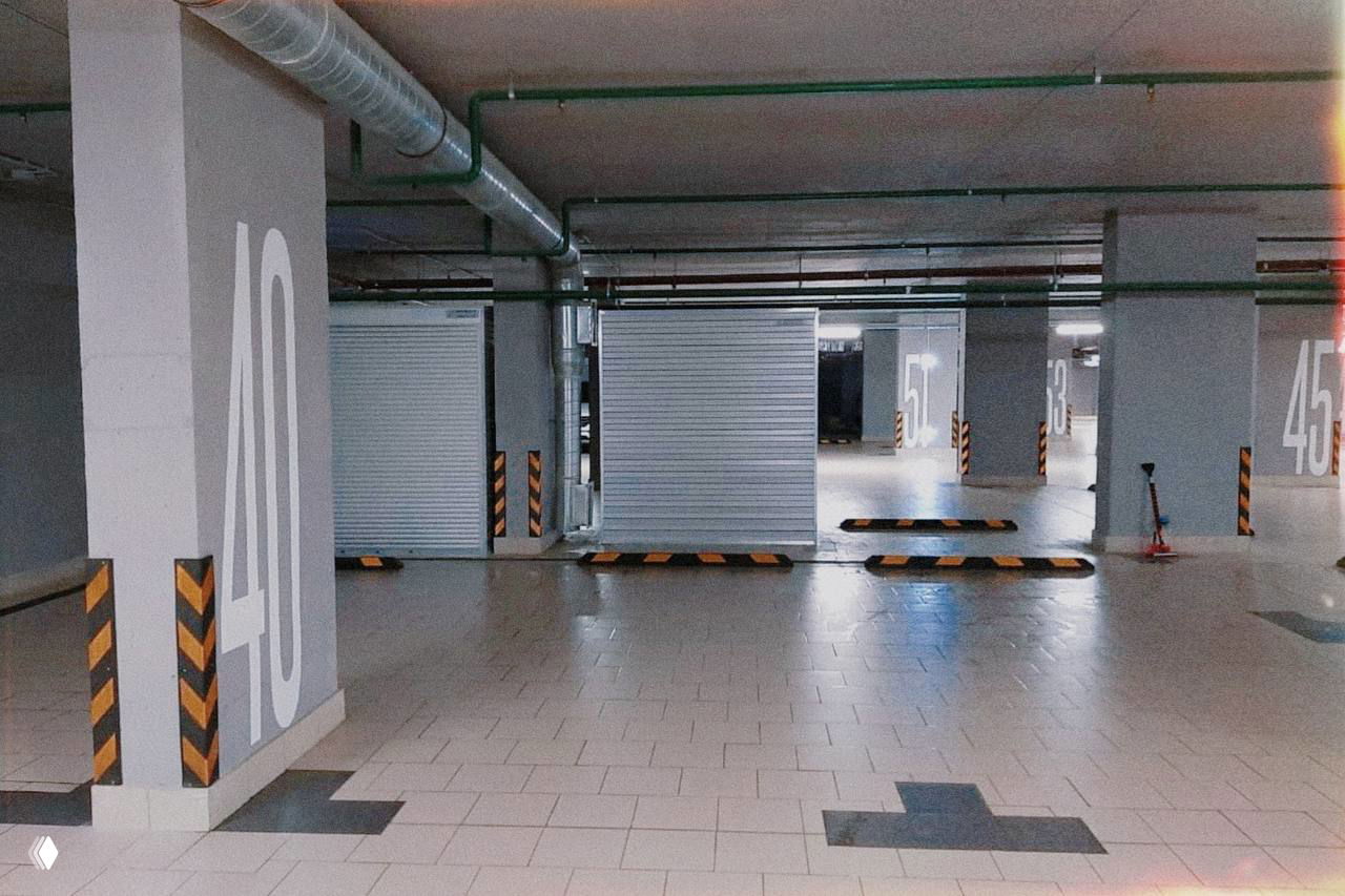 Вид подземного паркинга с размеченными машино-местами, плиточным полом, роллетами и номерами парковочных мест — пример продаваемого объекта.