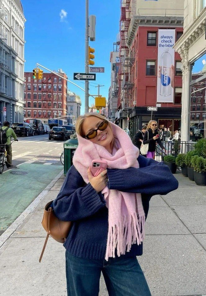 Улыбающаяся девушка на городской улице в тёмном свитере и большом розовом шарфе, держащая розовый телефон — динамичный стритстайл кадр.
