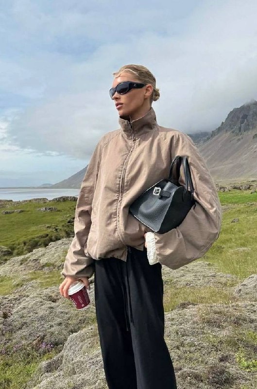 Уличный стиль: просторная бежево-песочная ветровка на фоне гор и озера, сумка и кофе в руке — практичный выбор для поездки за город.