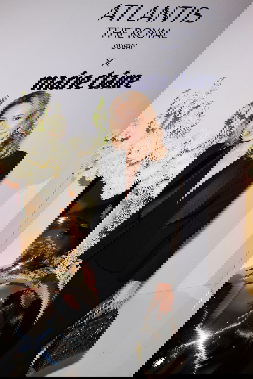 Гостья в белой блузке и чёрных брюках позирует у бренд-зоны Atlantis The Royal x Marie Claire на фоне мягкой цветочной композиции и отражающих шаров.