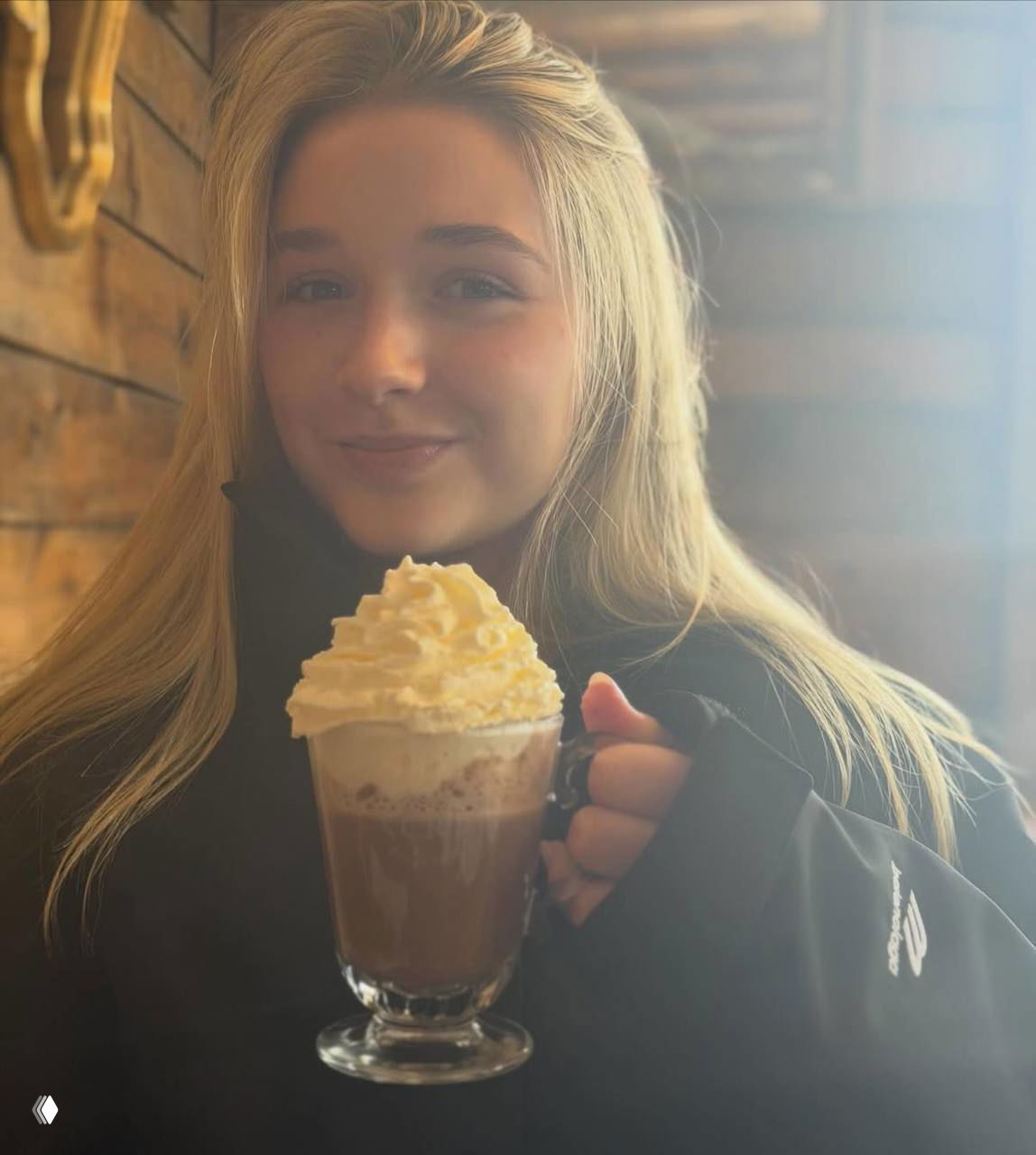 Девушка в тёплой одежде держит кружку горячего шоколада со взбитыми сливками в уютном интерьере горного шале.