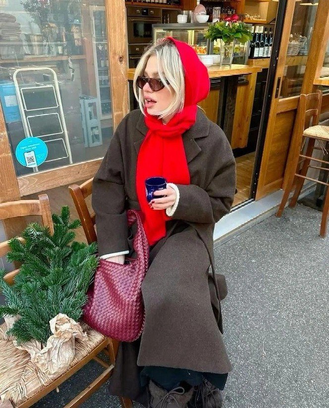 Женщина в ярком красном шарфе и капюшоне сидит в уличном кафе с кружкой в руках, цветовой акцент на шарфе и непринуждённая поза.