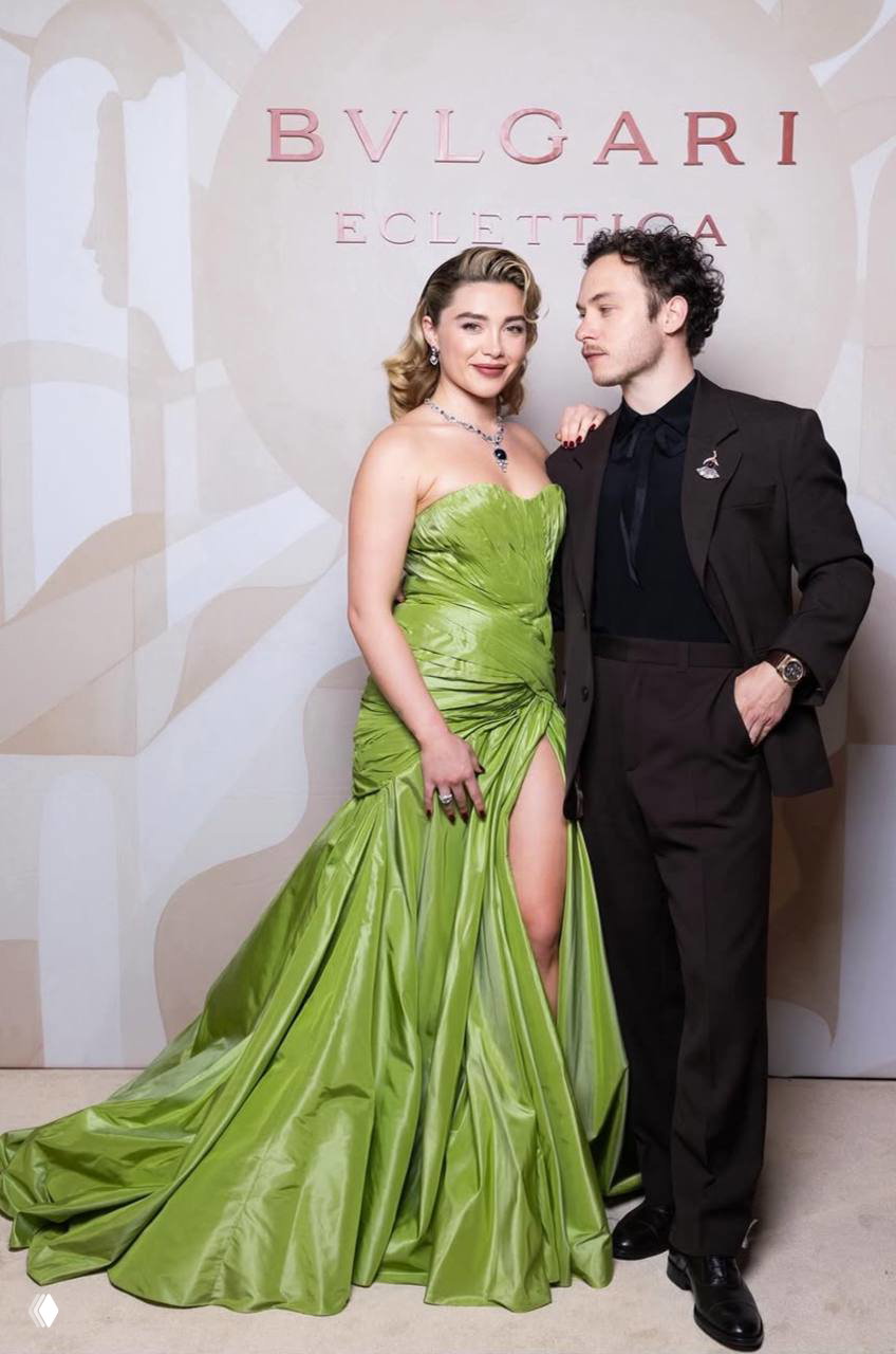 Женщина в ярком зелёном вечернем платье со шлейфом и мужчина в тёмном костюме позируют у фотозоны с логотипом Bulgari.