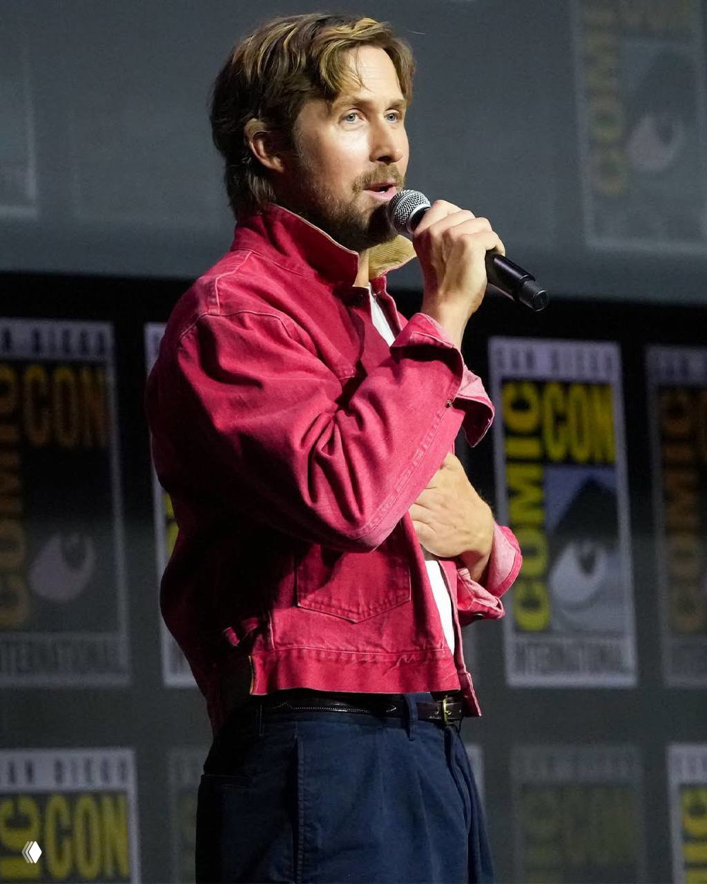 Крупный план актёра с микрофоном на Comic-Con: он в красной куртке, говорит со сцены, фон украшен баннерами мероприятия.