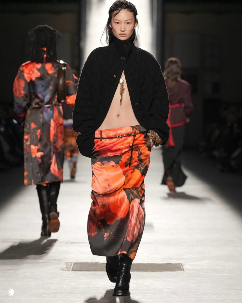 Модель в тёмной куртке и юбке с крупными оранжево-красными цветами, подчёркнутыми контрастом на подиуме Dries Van Noten.