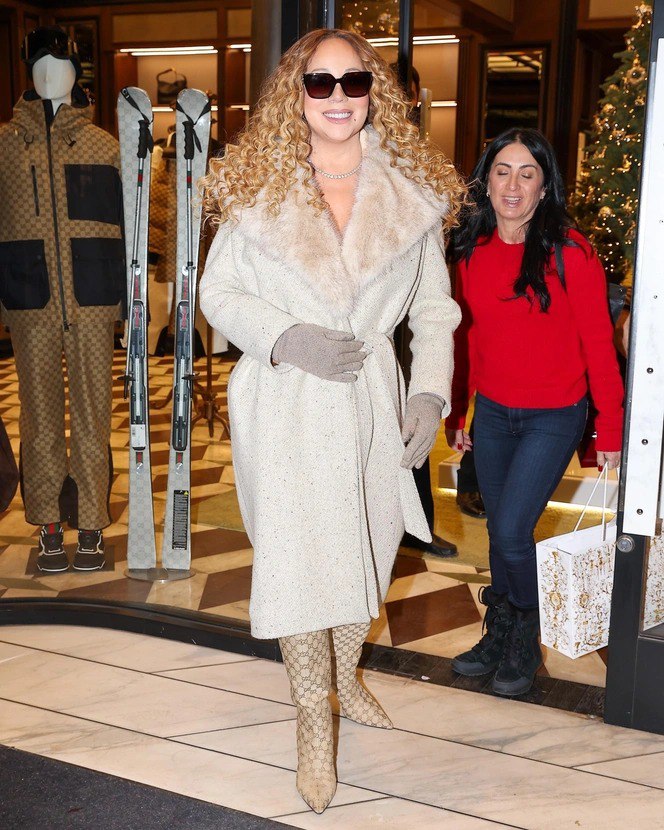 Певица в кремовом пальто‑халате с меховым воротником и перчатками, улыбается в солнцезащитных очках на фоне витрины бутика.