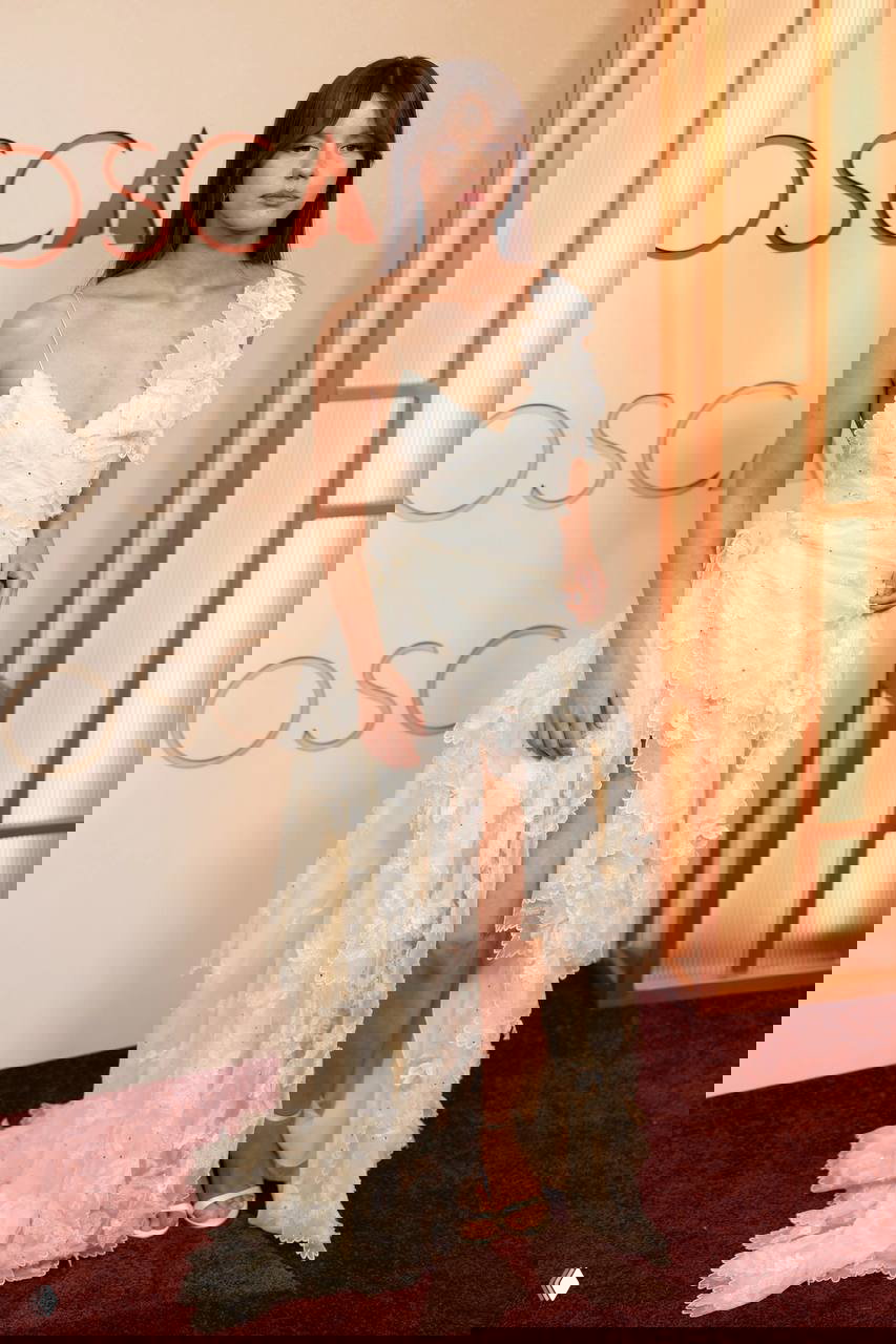 Модель в кремово-белом многослойном платье с кружевными и фактурными деталями, асимметричный декор и струящаяся юбка на красной дорожке.