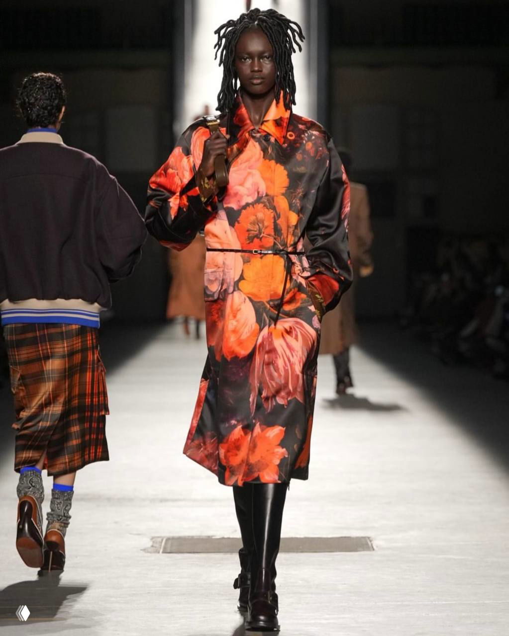 Пальто с ярким цветочным принтом шагает по подиуму — насыщенные оранжево-красные бутоны на тёмном фоне в сочетании с грубой обувью.