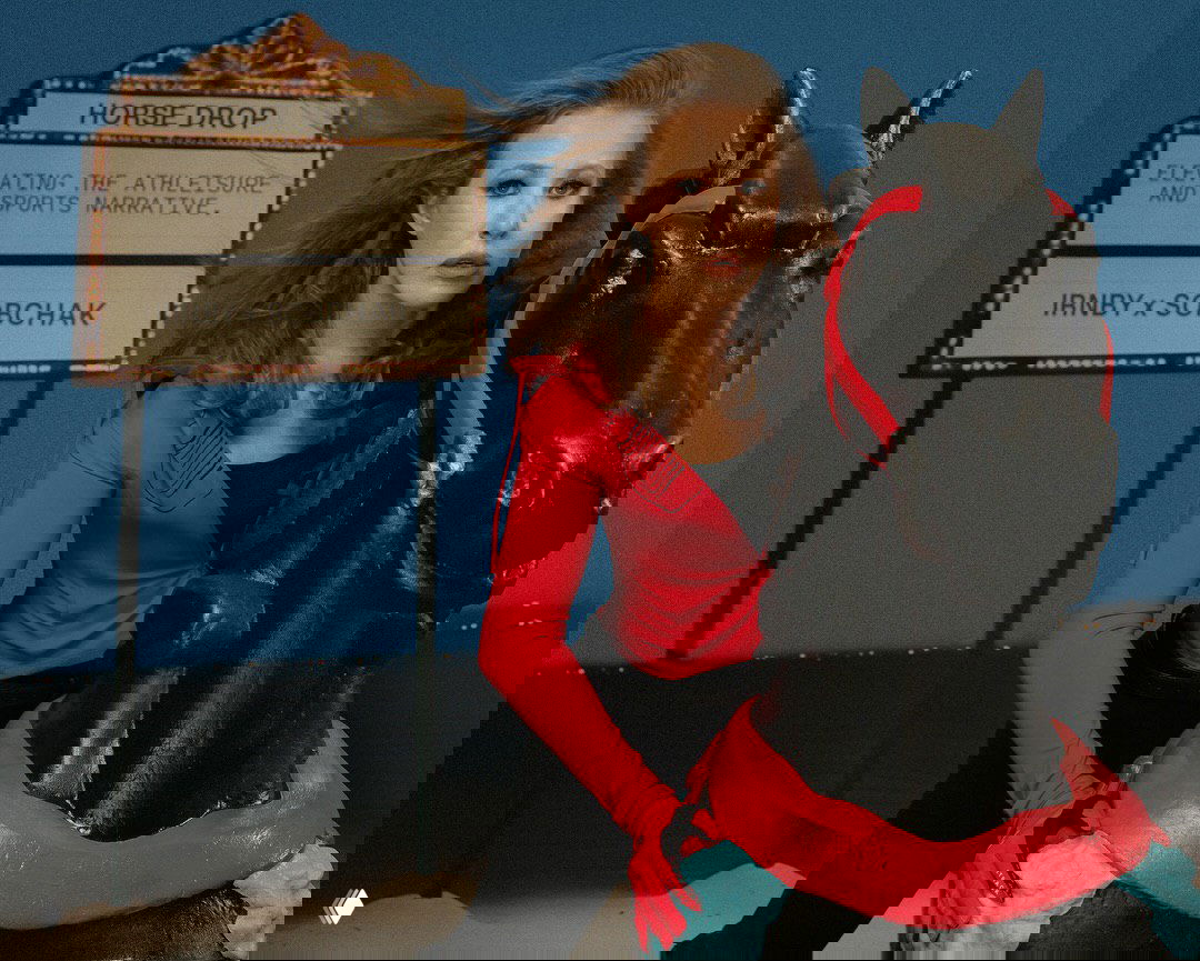 Женщина в красном спортивном верхе позирует рядом с чёрной декоративной статуей лошади и вывеской «HORSE DROP» — художественная съёмка для капсулы.