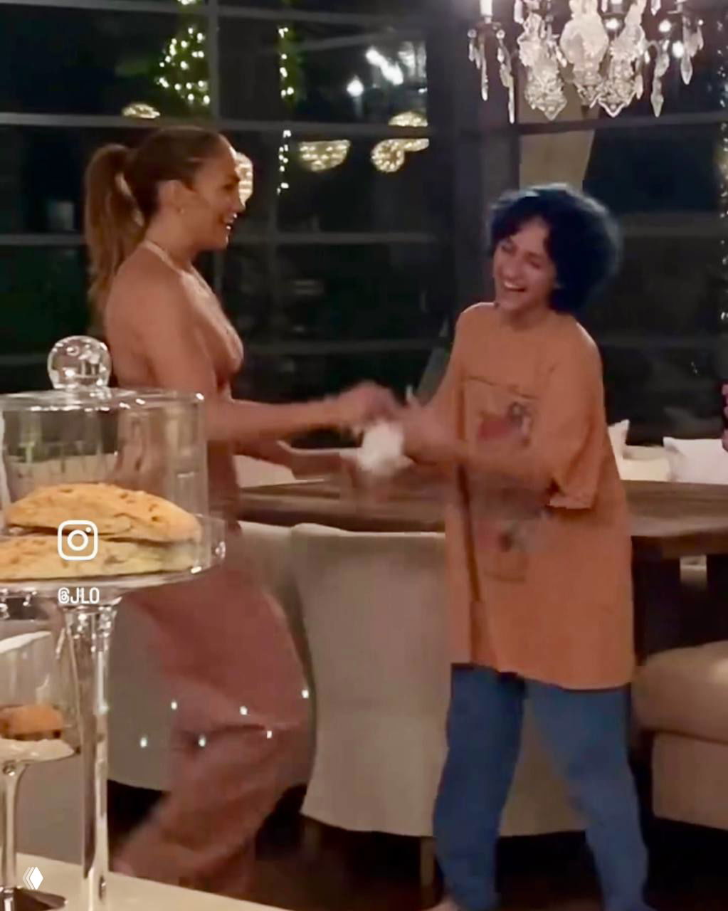 Две женщины и ребёнок смеются в столовой с люстрой и праздничным освещением; кадр с семейного праздничного ужина и радостной атмосферой