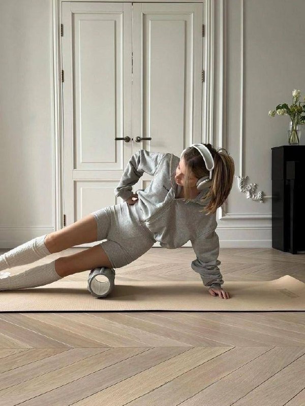 Женщина занимается с роликом для миофасциального массажа в светлой гостиной — кадр о восстановлении и работе с мышечным тонусом дома.