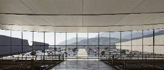 Вид на просторный интерьер павильона с панорамными окнами и видом на водную гладь и холмы — пространство, напоминающее павильон Serpentine Gallery.