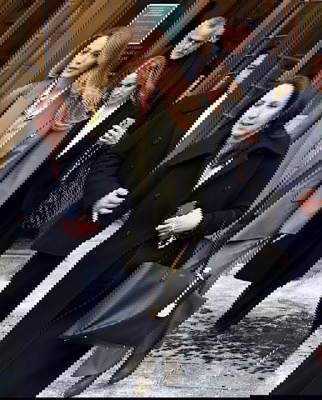 Группа людей покидает здание суда: женщина в оливковом костюме идёт впереди в окружении сопровождающих, папарацци снимают сцену с улицы.