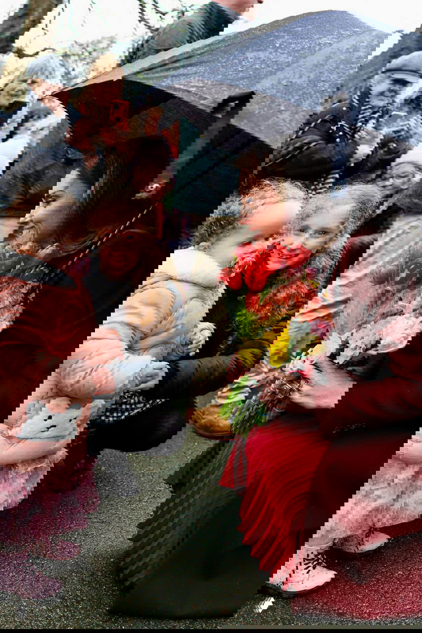 Женщина в бордовом пальто под зонтом принимает букет от маленькой девочки и улыбается на фоне толпы зрителей.