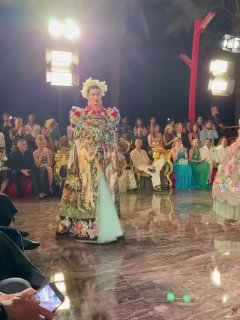 Модель в платье с богатой флористической аппликацией и длинным шлейфом идёт по зеркальному подиуму под светами вечернего показа