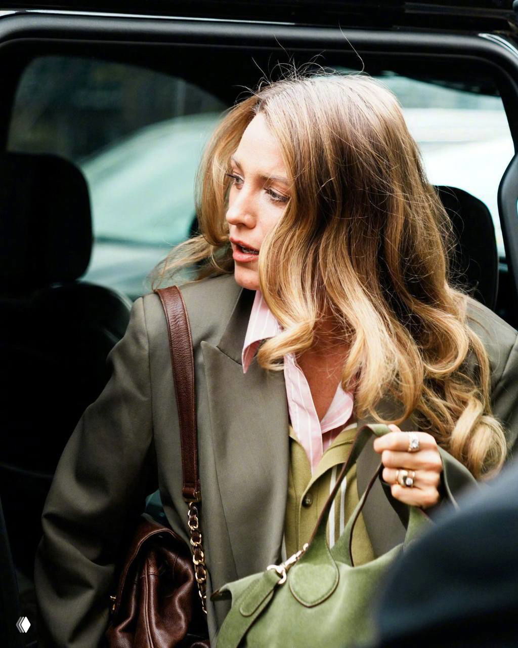 Крупный план женщины в светло-розовой рубашке и оливковой жилетке: детализация прически и сумки на фоне открытой дверцы автомобиля.