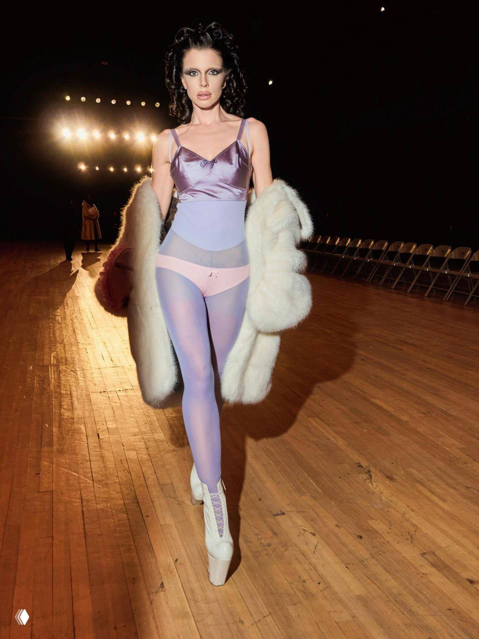 Модель идёт по подиуму в сиренево-розовом корсете, полупрозрачных колготках и белых высоких ботильонах на эстафетном деревянном полу сцены.