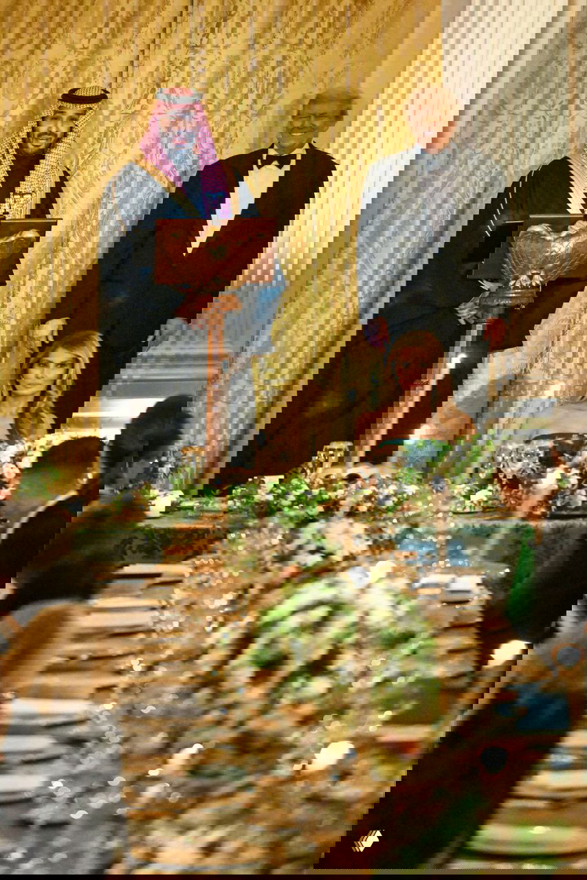 Стол с украшениями и рассаженными гостями на официальном банкете; в глубине кадра женщина в изумрудном платье сидит за столом во время выступления.