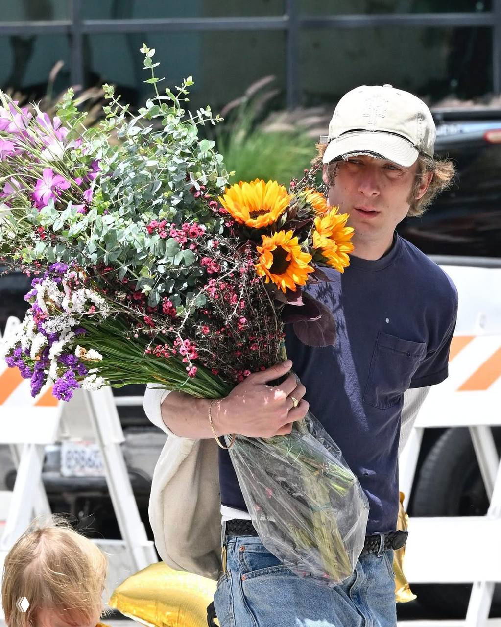 Крупный план: мужчина в кепке держит объёмный букет с подсолнухами и зелёными ветвями; видно упаковку цветов и дневной городской свет.