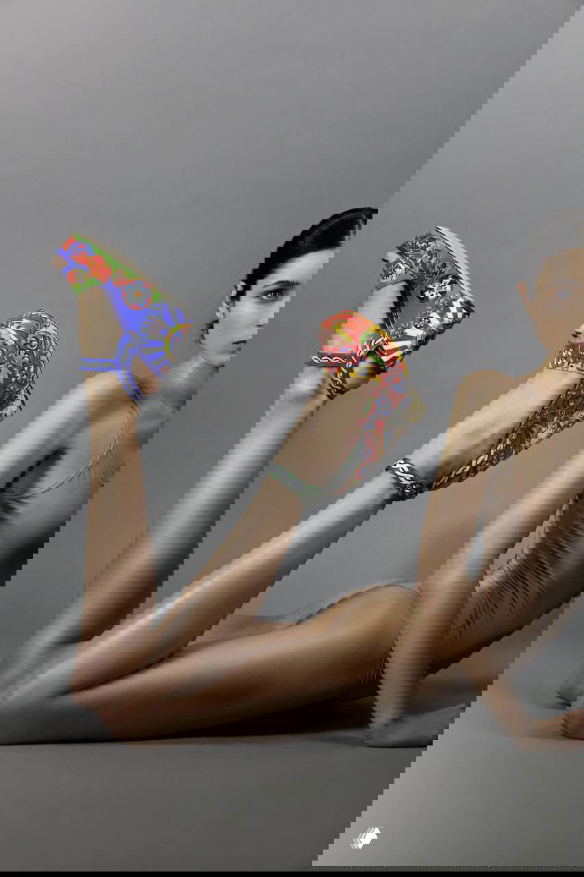 Модель в бежевом боди лежит на сером фоне, ноги подняты; акцент на яркие разноцветные танкетные босоножки с орнаментом.