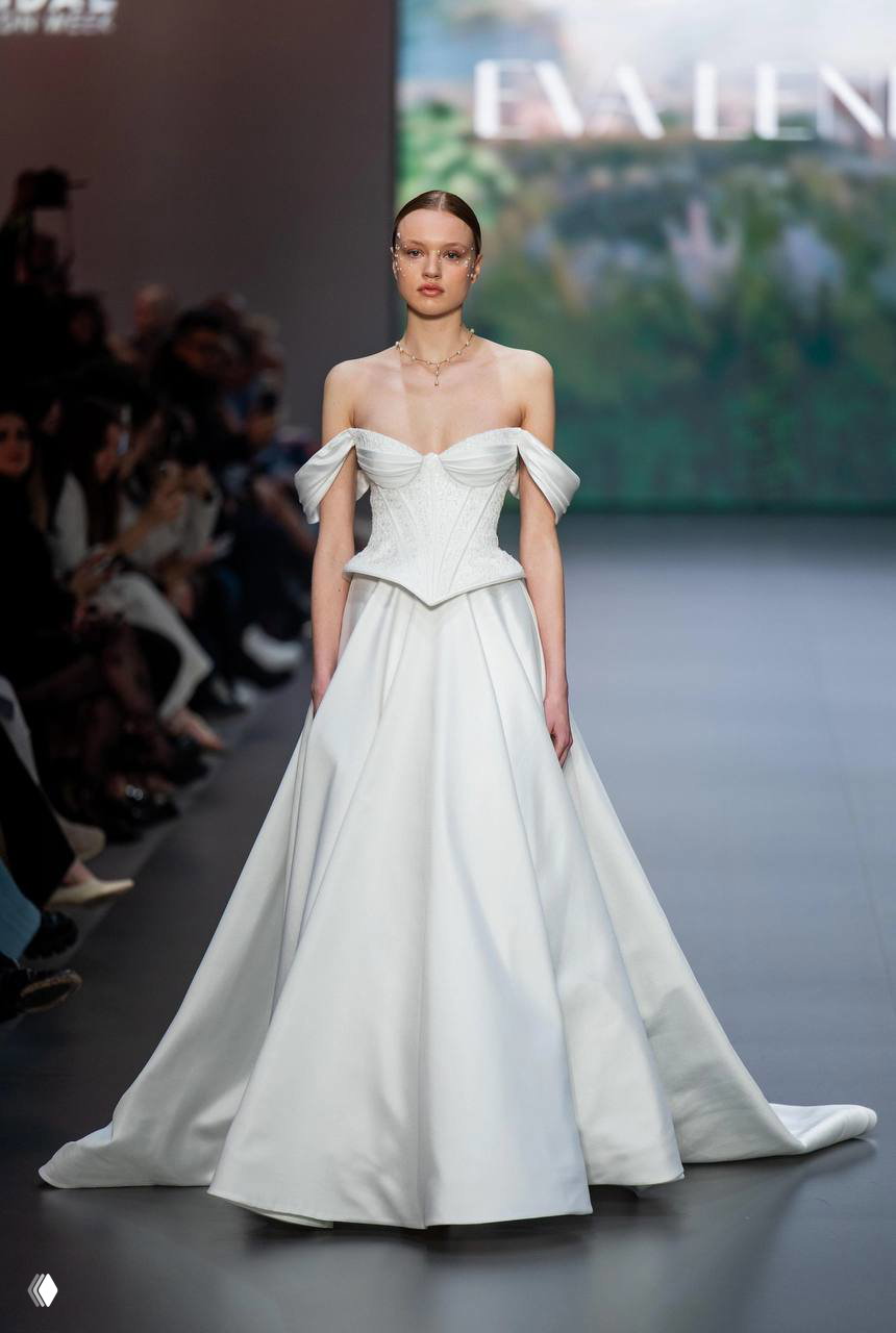 Модель в атласном свадебном платье с выраженным корсетом и открытыми плечами; строгая линия талии и мягкая юбка в показе мод.