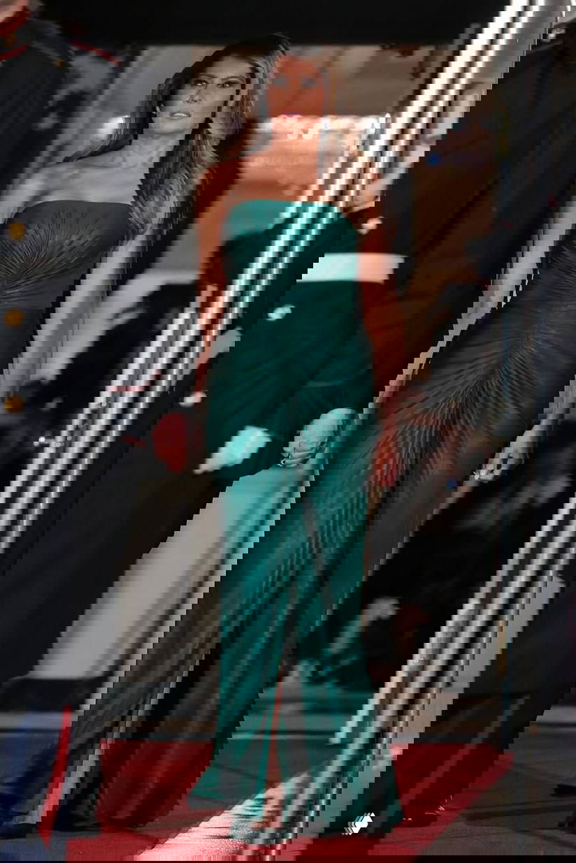 Женщина в изумрудном платье выходит через дверной проём по красной дорожке — длинный фасон с драпировкой и разрезом спускается к полу.