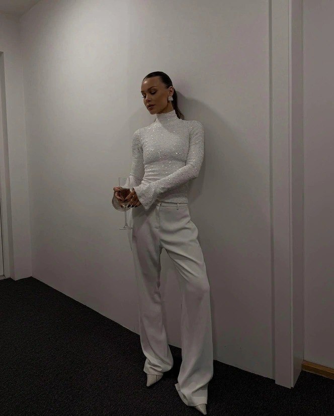 Модель в белых прямых брюках и блестящем светлом топе держит бокал, строгая поза на нейтральном фоне — идея праздничного образа с белыми брюками.