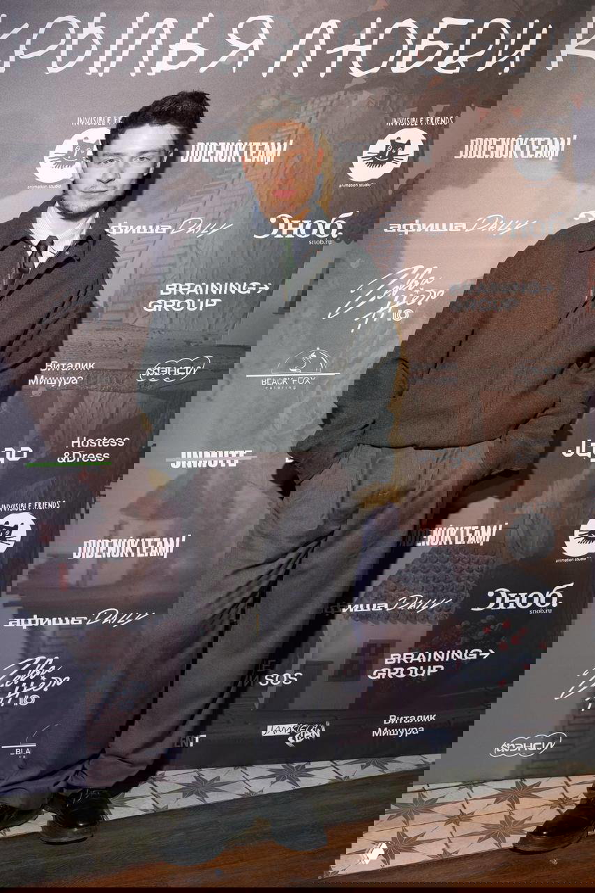 Посетитель в строгом образе стоит у фотозоны премьеры «Крылья любви», видно название фильма и бренды партнёров на заднем плане.