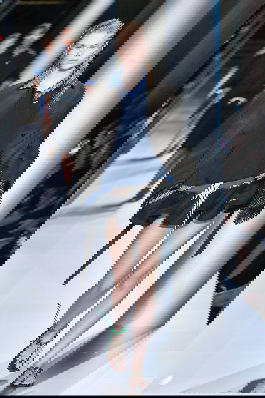 Модель на подиуме в мини-юбке, гетрах и объемном пиджаке — образ в духе Miu Miu, показ с небрежной прической и аксессуарами.