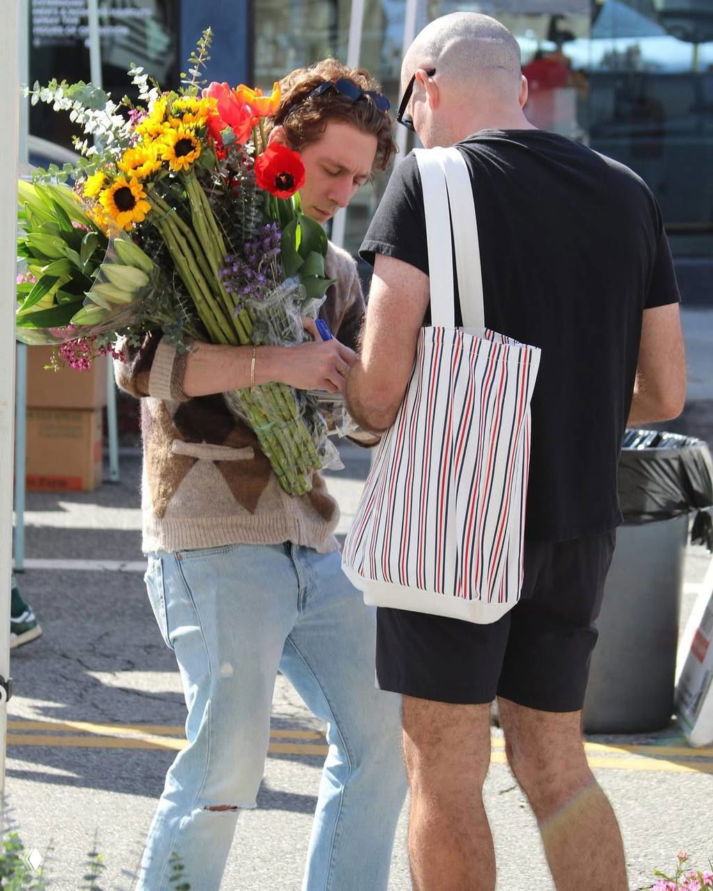 Мужчина подписывает что-то, одновременно держа большой пышный букет с подсолнухами и разноцветными летними цветами; уличная сцена взаимодействия с прохожими.