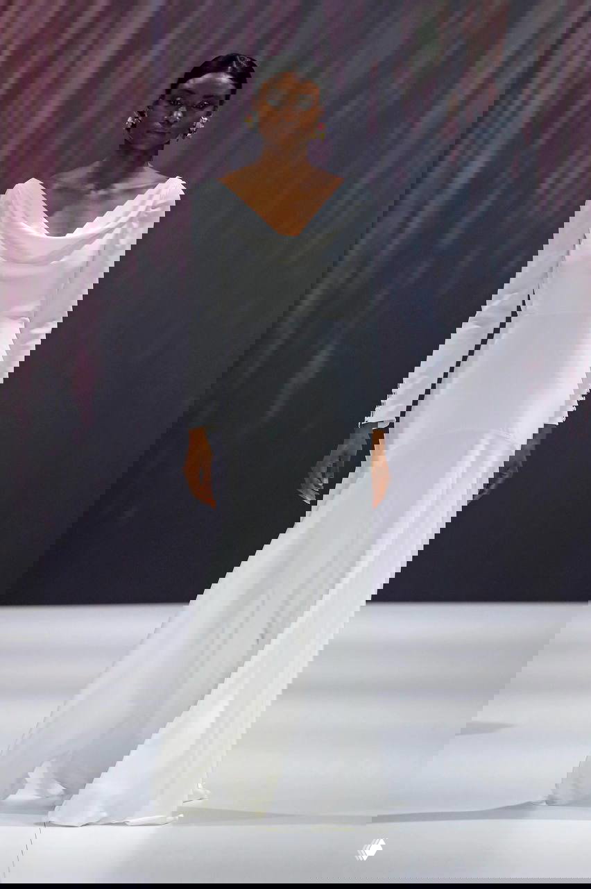 Модель в белом платье со свободным драпированием и длинными рукавами; минималистичный силуэт и плавная фактура ткани на подиуме.