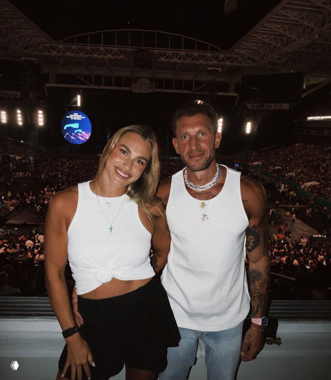 Арина Соболенко и Георгиос Франгулис позируют вместе в зрительном зале стадиона, пара улыбается на фоне большого концерта или события.